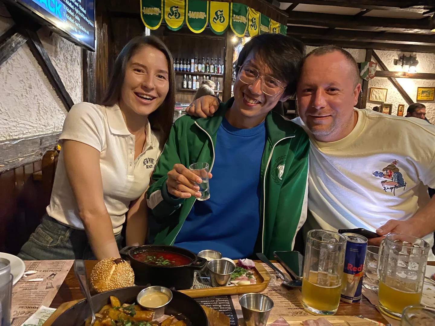 三个人坐在餐桌前,中间的人穿着绿色外套和蓝色内搭,手里拿着一杯透明饮料。左边的人穿着白色短袖衬衫,右边的人穿着印有图案的白色T恤。餐桌上摆放着食物、饮料和餐具,背景中有酒瓶和装饰旗帜。