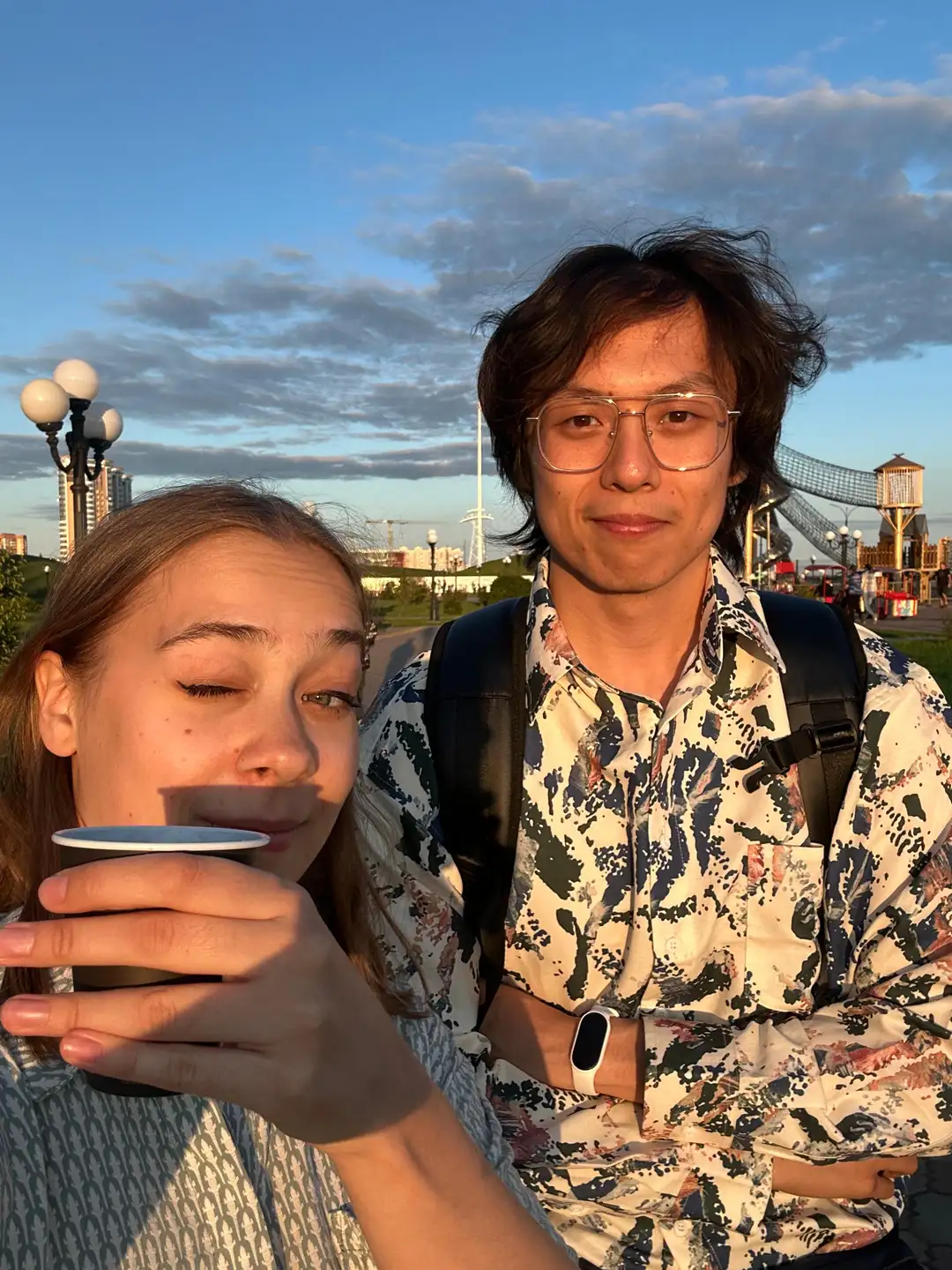 两个人在户外合影,其中一人手持杯子正在喝东西,背景有灯光柱和游乐设施。天空呈现蓝色并带有云彩,远处可见建筑物。