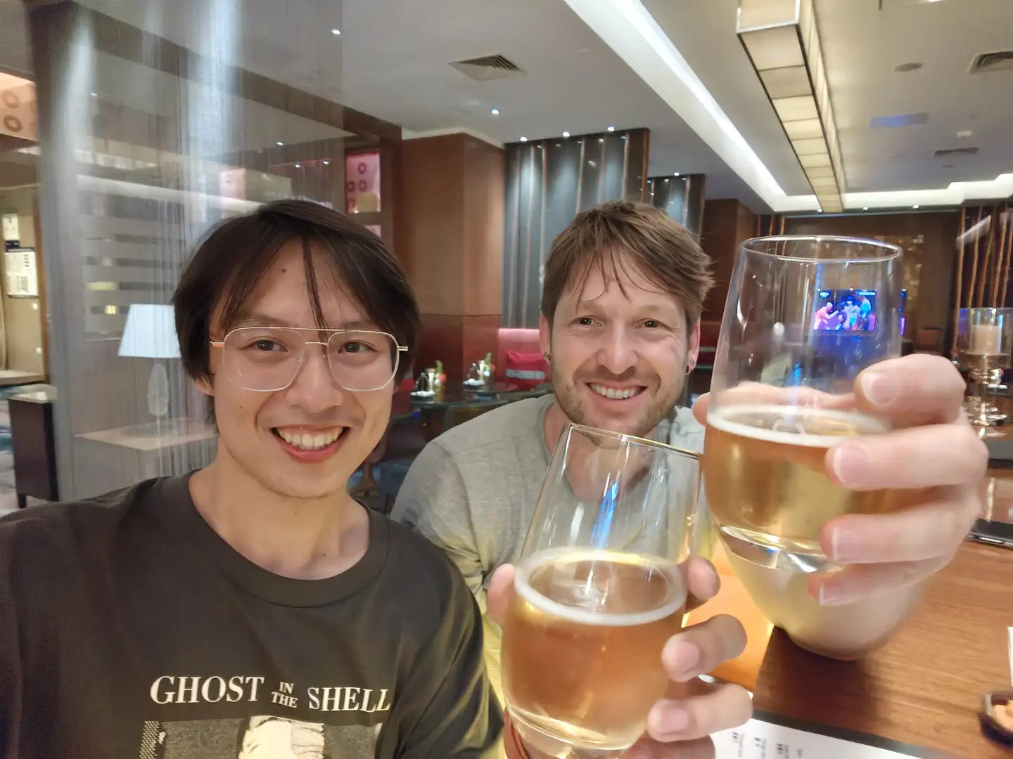 两个人举着啤酒杯,杯子中有浅色液体,背景是一个室内环境,装饰现代。他们穿着休闲服装,一人穿灰色T恤,另一人穿印有“GHOST IN THE SHELL”字样的黑色T恤。背景中可以看到一些餐厅设施和装饰。