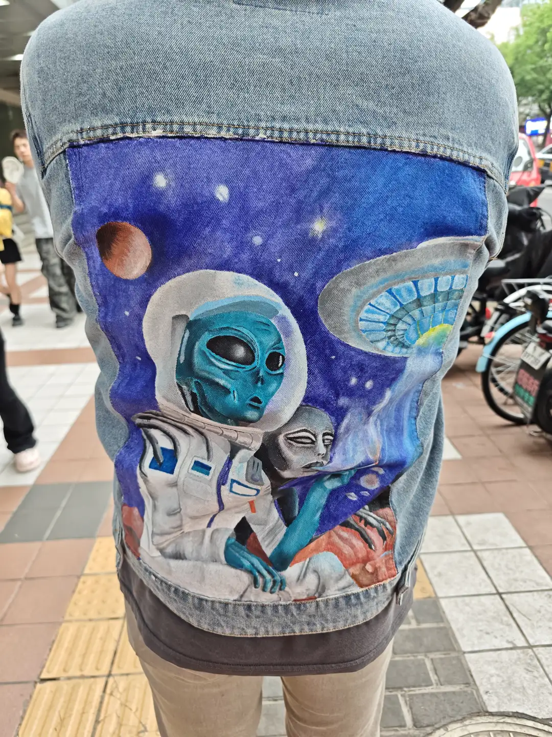 一个人穿着牛仔夹克,夹克背面绘有太空人和外星人的图案。背景是街道场景,地面铺有砖块。旁边有行人和自行车。