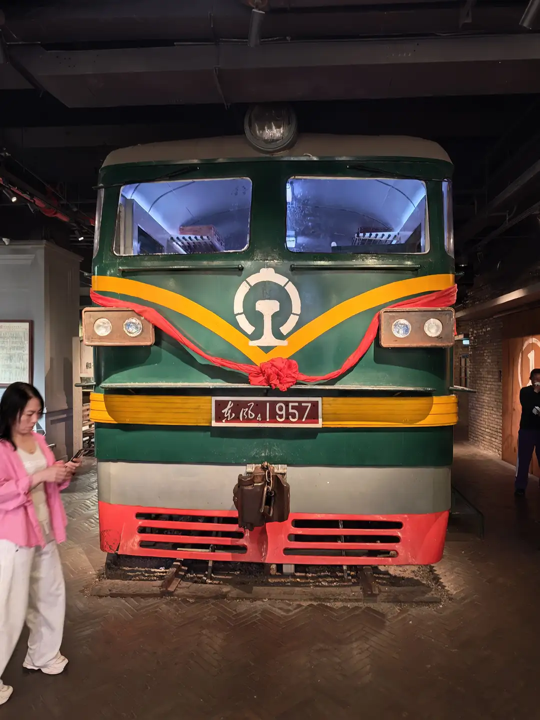 一辆绿色火车头停在室内展览空间,车头前系着红色绸带。火车编号为东风4 1957,前方有圆形标志。一名女子站在火车旁看手机。
