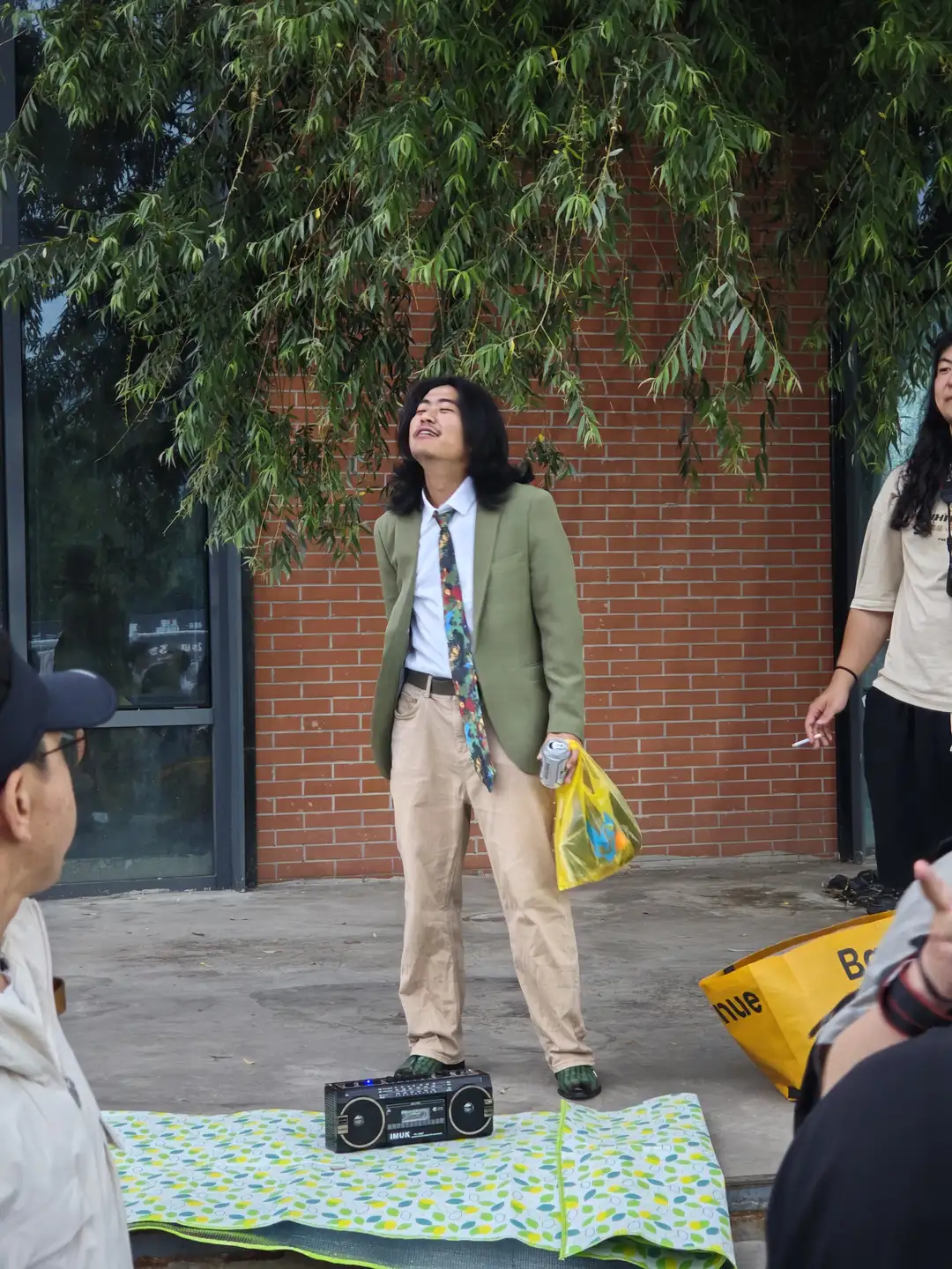 一名男子站在户外,手持塑料袋和罐装饮料,旁边放置一台老式收音机。男子穿着绿色西装外套和卡其裤,背景为红砖墙和垂柳。旁边有几个人在观看。