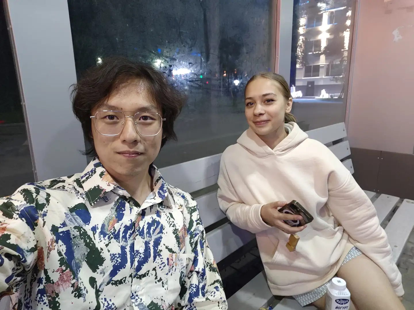 两个人坐在长椅上,左边的人穿着花衬衫,右边的人穿着浅粉色连帽衫,手里拿着手机,旁边放着一瓶酸奶,背景是夜晚的玻璃窗。