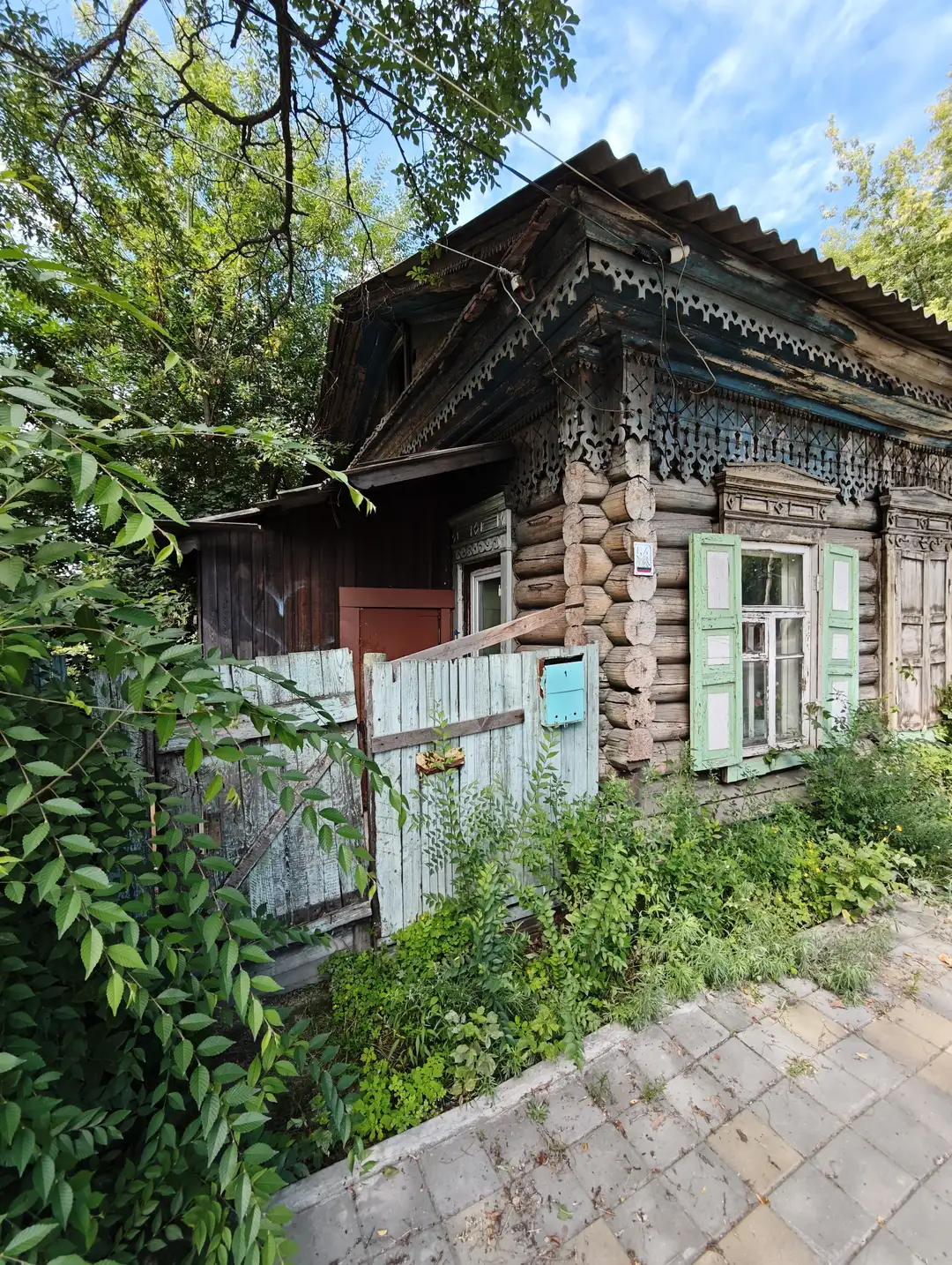 木制小屋由原木搭建,屋顶为波纹铁皮,窗户带有绿色百叶窗,门前有低矮的木栅栏和茂密的绿植。房屋部分结构陈旧,墙角有雕刻装饰,周围树木环绕,环境宁静。