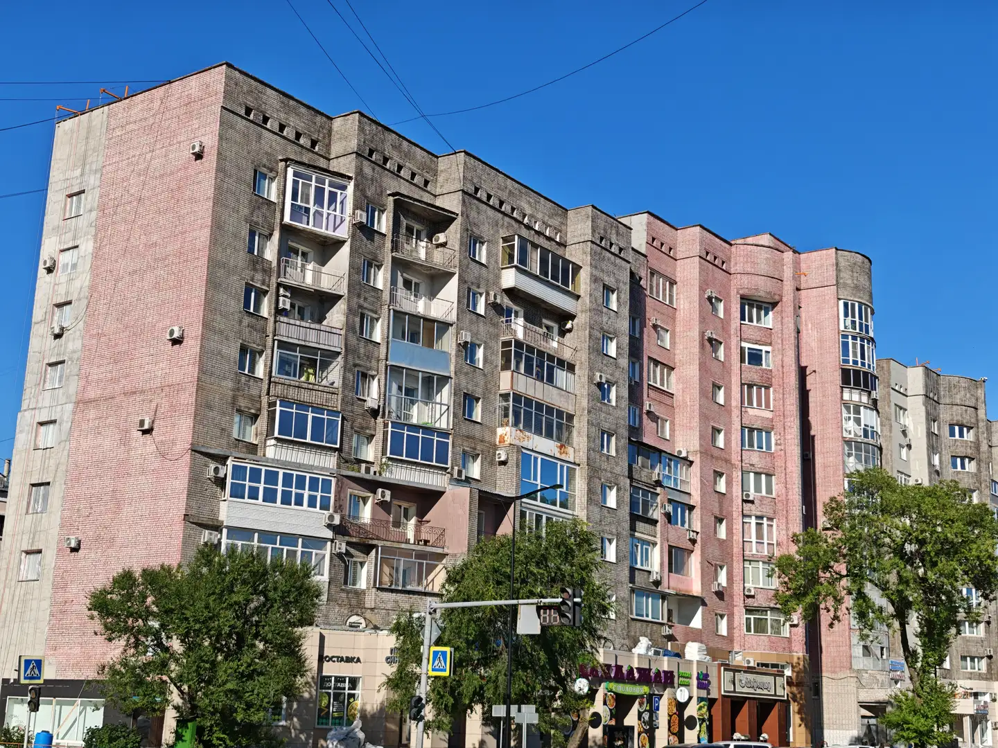 一栋多层住宅楼,外立面由粉色和灰色砖块构成,窗户排列整齐,部分阳台装有玻璃窗或金属栏杆。楼体底部设有商铺,招牌上有“ОCTABKA”等字样,门前有交通信号灯和行人过街标志。周围有绿树和电线,天空晴朗无云。