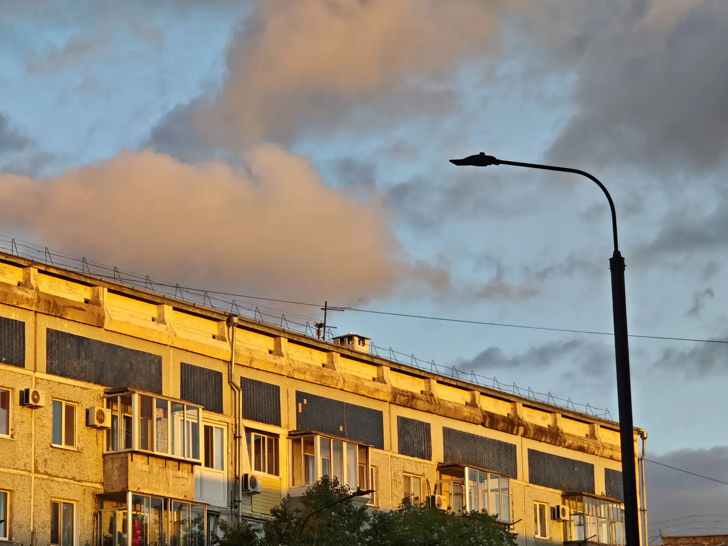 一排老旧住宅楼的侧立面被夕阳染成暖黄色,屋顶上设有金属围栏和小型通风口。窗户多数为封闭式,部分带有玻璃阳台,外墙上安装多个空调外机。右侧有一根高耸的路灯杆,背景是蓝灰色的天空和蓬松的云层。