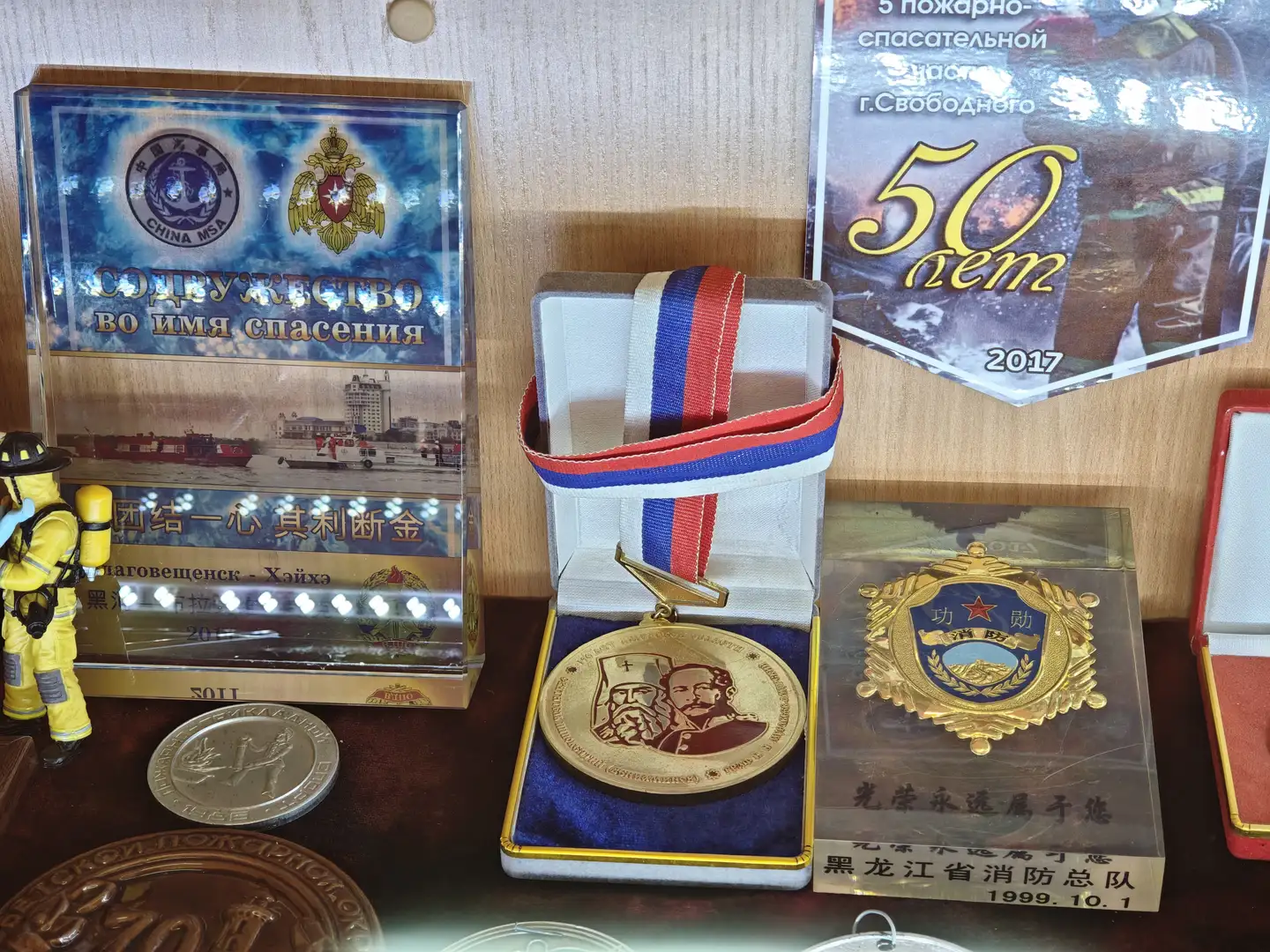 展台上摆放着一枚金色奖章,奖章上刻有两个人物肖像,系着红蓝白三色绶带。左侧是一个透明展示盒,内有消防主题的纪念品和中文标语“团结一心 其利断金”。右侧是一个金色徽章,下方有中文文字“黑龙江省消防总队 1999.10.1”,旁边还有一张庆祝50周年的海报。