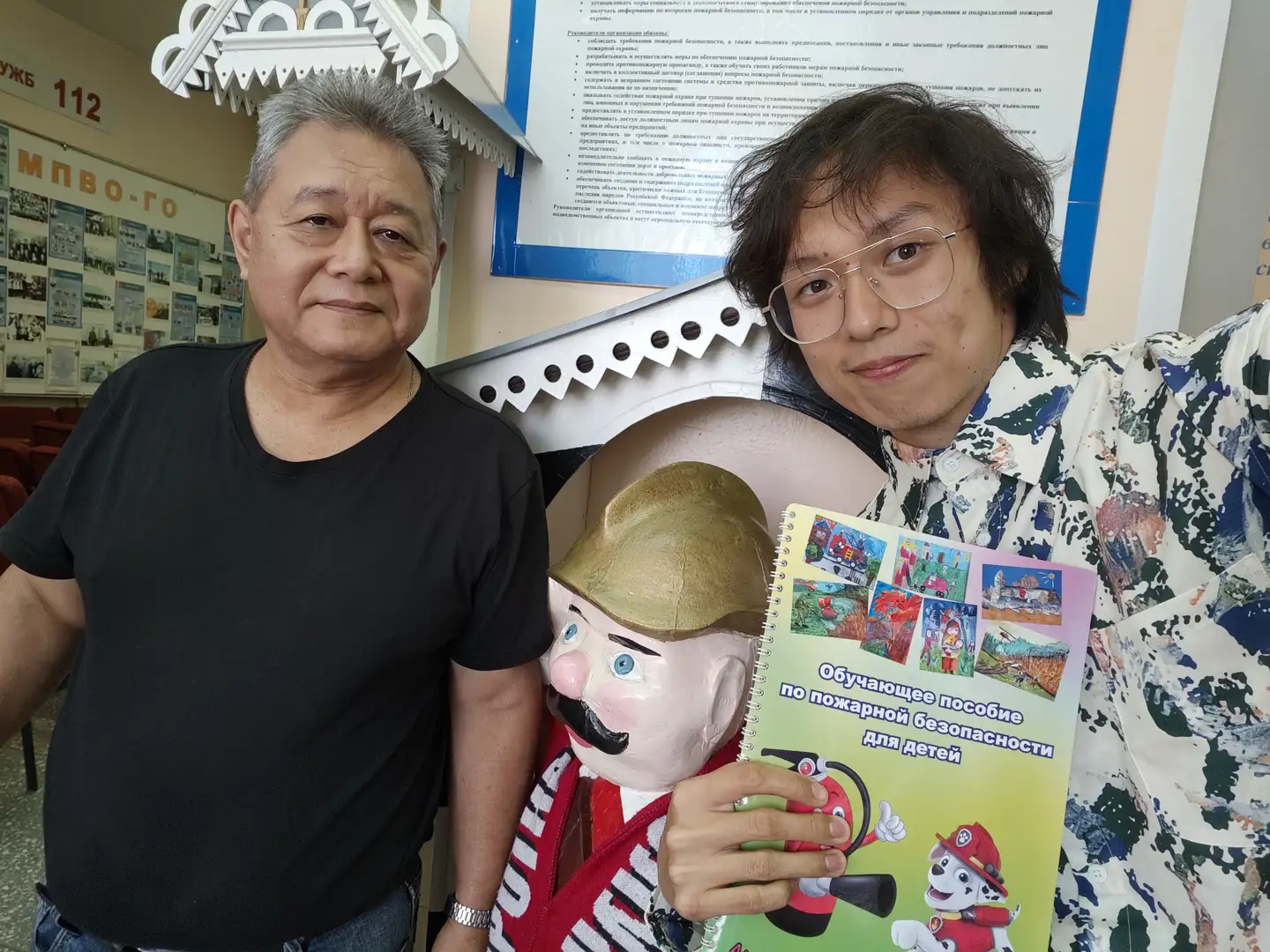 两位男士站在一个卡通消防员雕像前合影,其中一人手持一本封面印有消防狗图案的彩色儿童读物。背景墙上贴有消防知识宣传资料,上方有数字“112”和俄文标识。两人面带微笑,身后环境为室内,有装饰性屋顶结构。