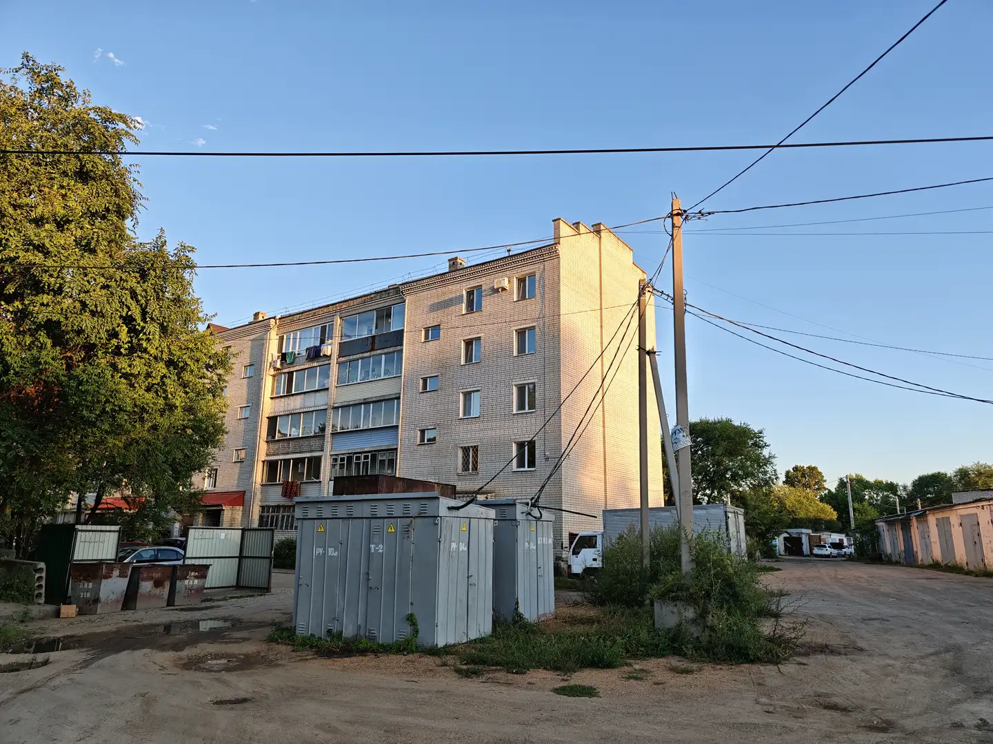 一栋五层高的住宅楼矗立在画面中央,外墙为浅色砖块,窗户排列整齐。楼前有几台灰色的电气箱和一些低矮的建筑。右侧有一根电线杆,天空晴朗,阳光洒在建筑物上。