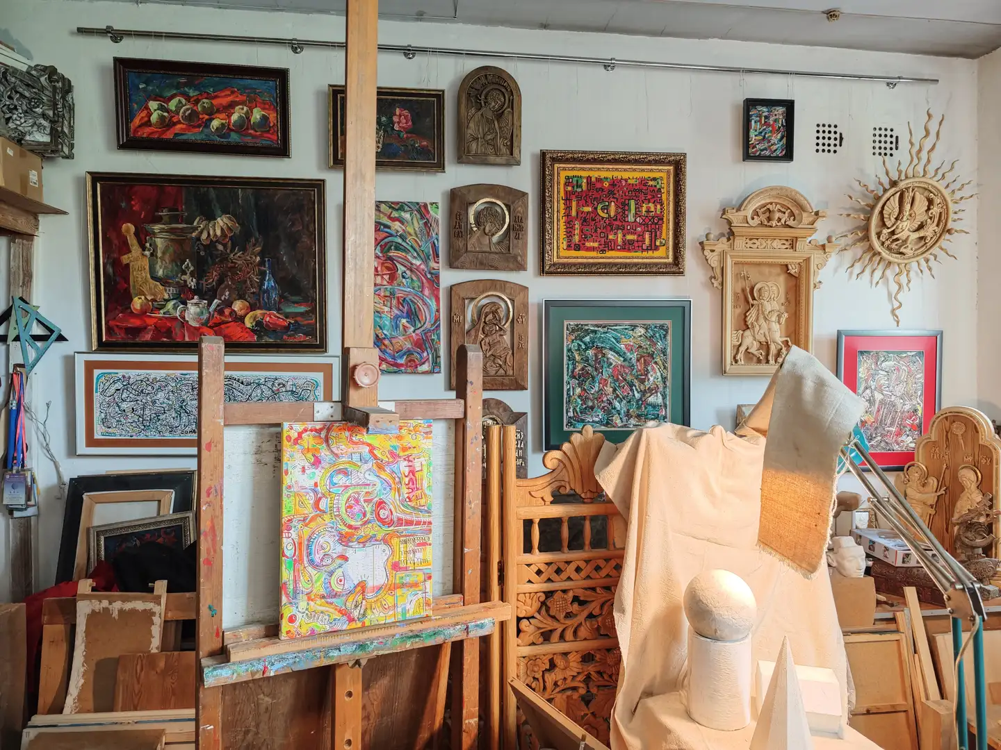 画室墙壁上挂满了各种艺术作品,包括静物画、宗教浮雕、抽象画和装饰性木雕。房间中央有一个画架,上面放置着一幅色彩丰富的抽象画,旁边是一个覆盖布的模特台,上面摆放着几何形体。地面和角落堆放着画框和木制品,环境显得充实而富有创作气息。
