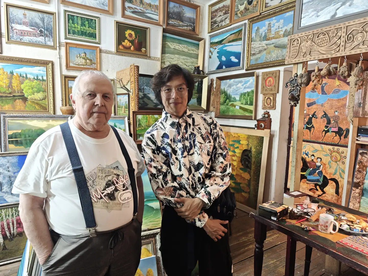一位年长的男士和一位年轻男子站在一间挂满风景画的房间里,墙上陈列着许多装裱好的画作。年长者穿着白色T恤和背带裤,年轻男子穿着带有复杂图案的衬衫和黑色裤子。房间的一角有一张桌子,上面放着一些物品,包括一个杯子和食物。