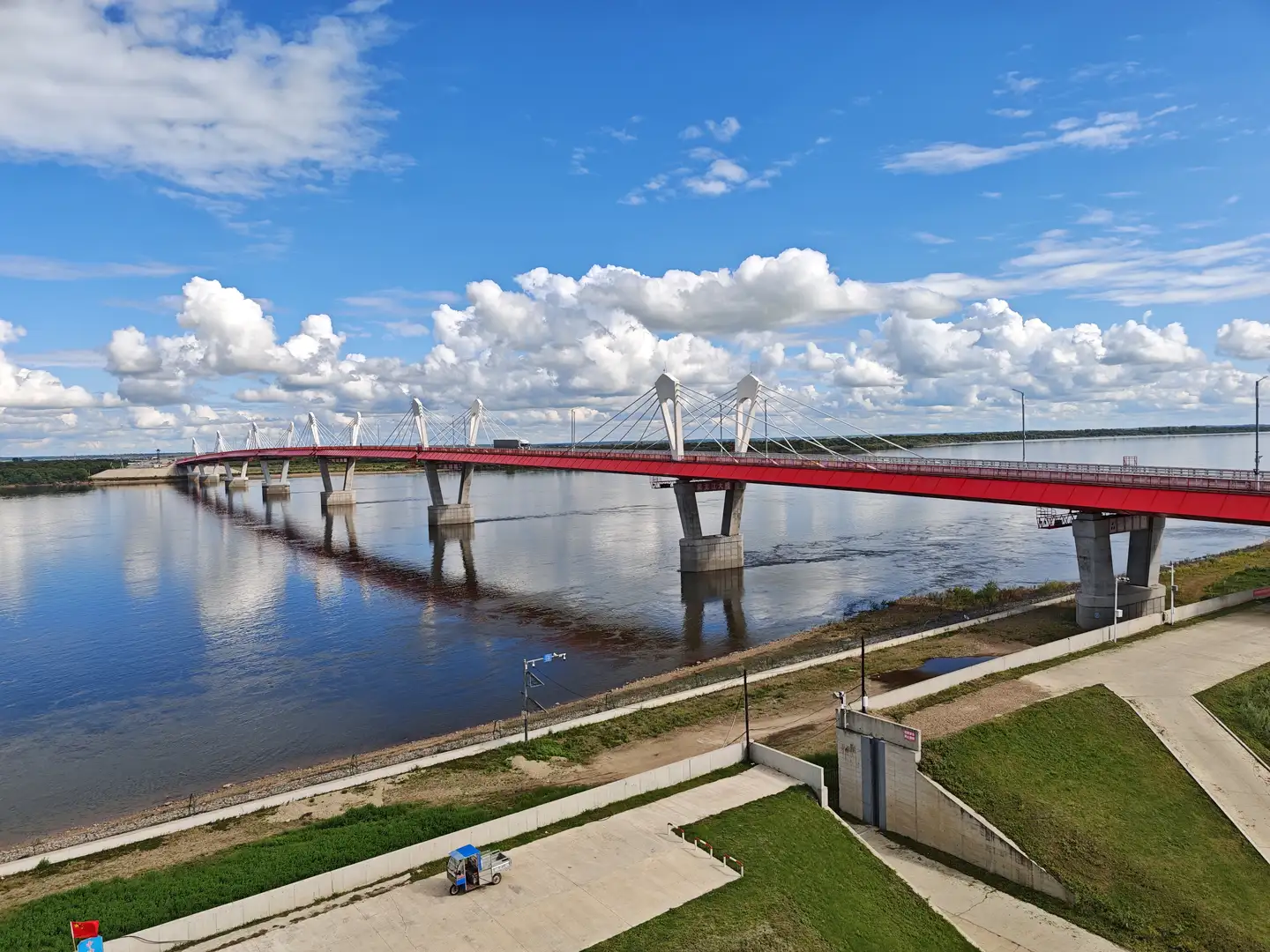 一条宽阔的河流上横跨着一座红色的斜拉桥,桥身由多个混凝土桥墩支撑。河岸两侧有绿化带和小路,岸边停着一辆蓝色顶棚的小车。天空湛蓝,布满蓬松的白色云朵。