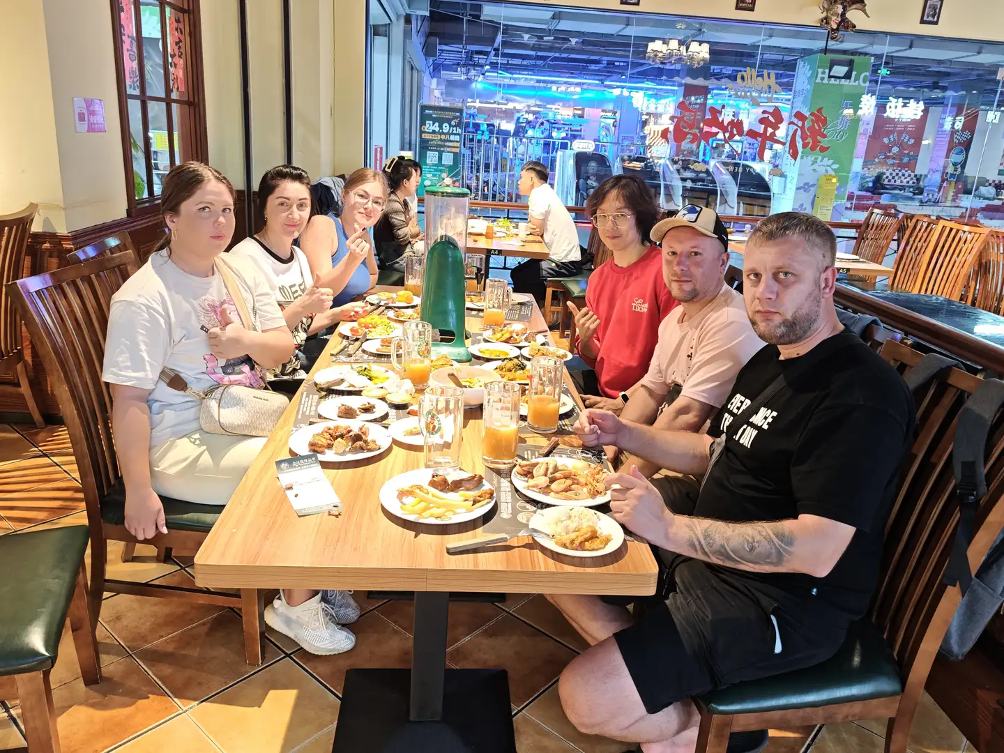 一群人围坐在餐厅的长桌旁,桌上摆满了食物和饮料,包括薯条、肉类和橙汁。他们穿着休闲服装,有的面带微笑,有的做出手势,背景中可以看到其他顾客和店铺招牌。餐厅内部装饰温馨,有木质桌椅和玻璃窗,窗外是明亮的商业街景。
