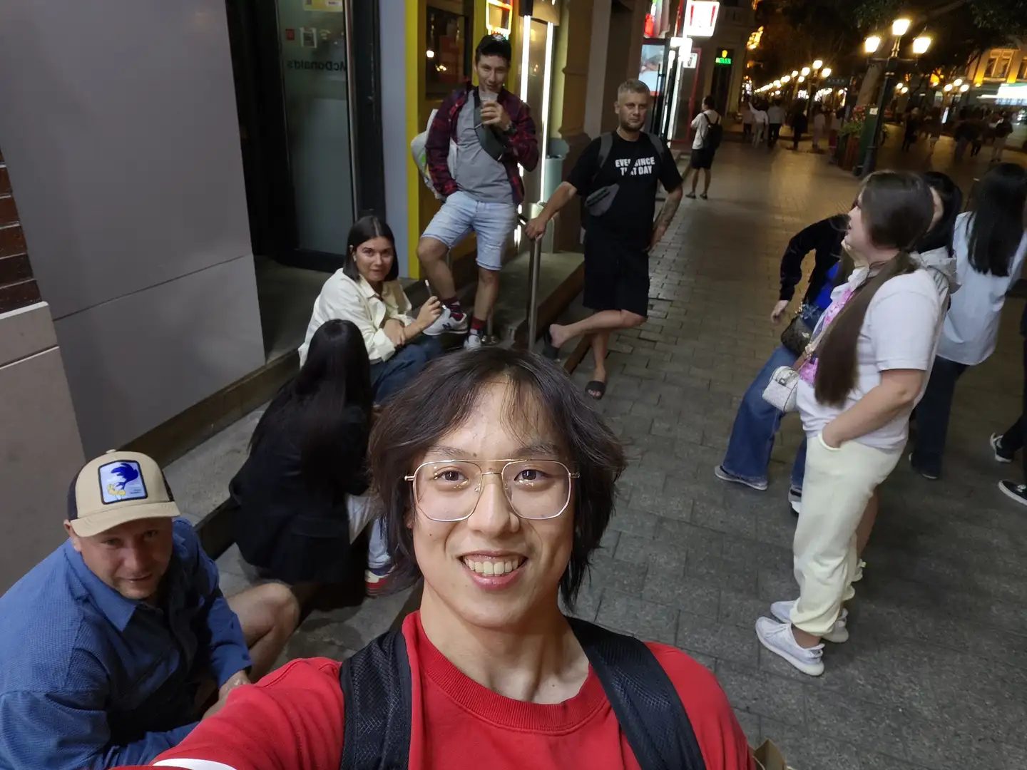 一名戴眼镜的男子在街道上自拍,他穿着红色上衣背着黑色背包,周围有多人站立或坐着。背景是夜晚的城市街道,灯光照亮了行人和商店,地面铺有石砖。人们穿着夏季服装,有的在交谈,有的在看手机。