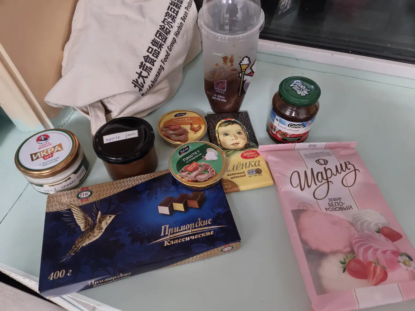 桌面上摆放着多种食品,包括一盒印有飞鸟图案的巧克力、一袋粉色包装的糖果、几罐果酱和肉制品、一杯奶茶和一个印有中文的布袋。巧克力包装上有俄文“Приморские Классические”,糖果包装上写有“Марица”和“Зефир Бело-Розовый”。旁边还有标有“apple jam”的罐子和带有“Global Village”字样的果酱瓶。