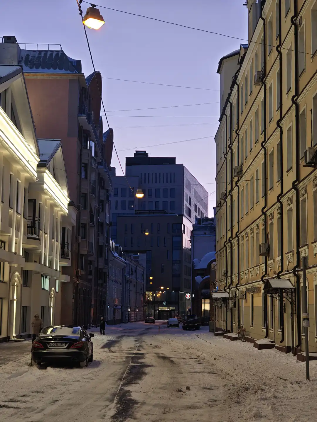 拂晓的街道,莫斯科 一条覆盖着积雪的街道延伸向远方,两侧矗立着高低错落的建筑。左侧是一栋带有明亮灯光装饰的白色现代建筑,旁边停着一辆车牌号为X001YX 777的黑色轿车,右侧则是一栋多层黄色老式建筑,外墙上有黑色的排水管。街道尽头可以看到几栋深色的现代高楼,天空中悬挂着几盏发出暖黄色光芒的路灯,路面留有车辆行驶过的车辙印。