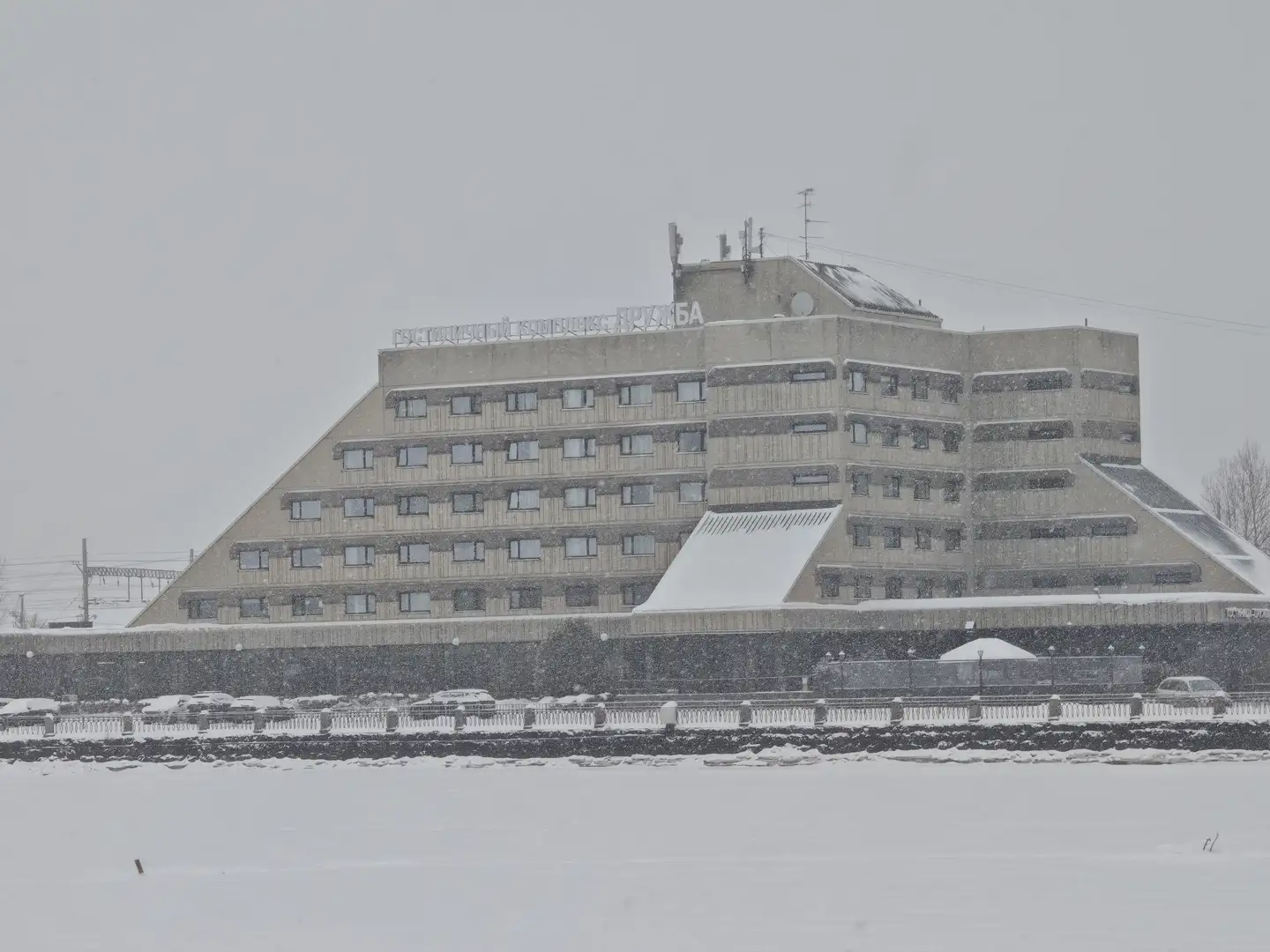 白雪覆盖的混凝土大楼,维堡 这座宏伟的阶梯状混凝土建筑拥有三角形的轮廓,层层叠叠的阳台结构使其看起来像古代的金字塔。建筑顶部安装有天线和通讯设备,外墙上排列着许多整齐的矩形窗户。此时正值大雪纷飞,灰色的天空中飘洒着密集的雪花,地面和建筑表面都覆盖了一层白雪。