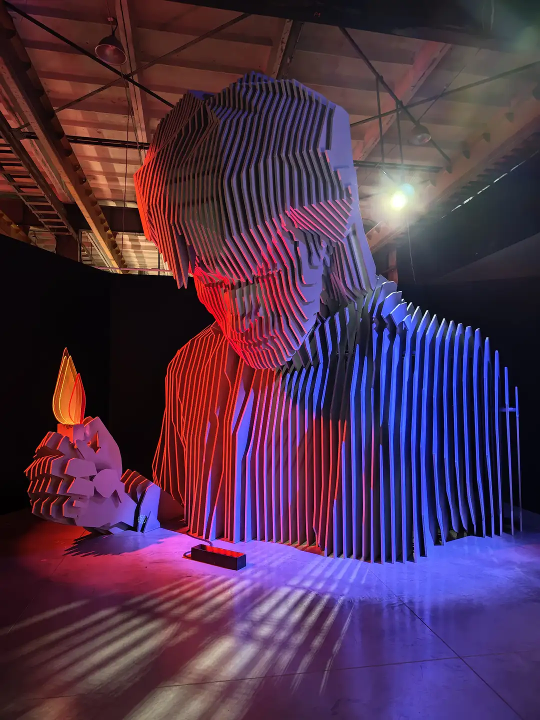 维克多·崔的主题展览,圣彼得堡 一个巨大的现代主义雕塑矗立在昏暗的室内空间,该雕塑由层层叠加的垂直板材构成,呈现出一个人物低头沉思的侧面轮廓。雕塑被红色和蓝色的灯光照射,使其边缘呈现出鲜明的色彩对比。人物雕塑的左手托举着一个类似火焰形状的发光物体。