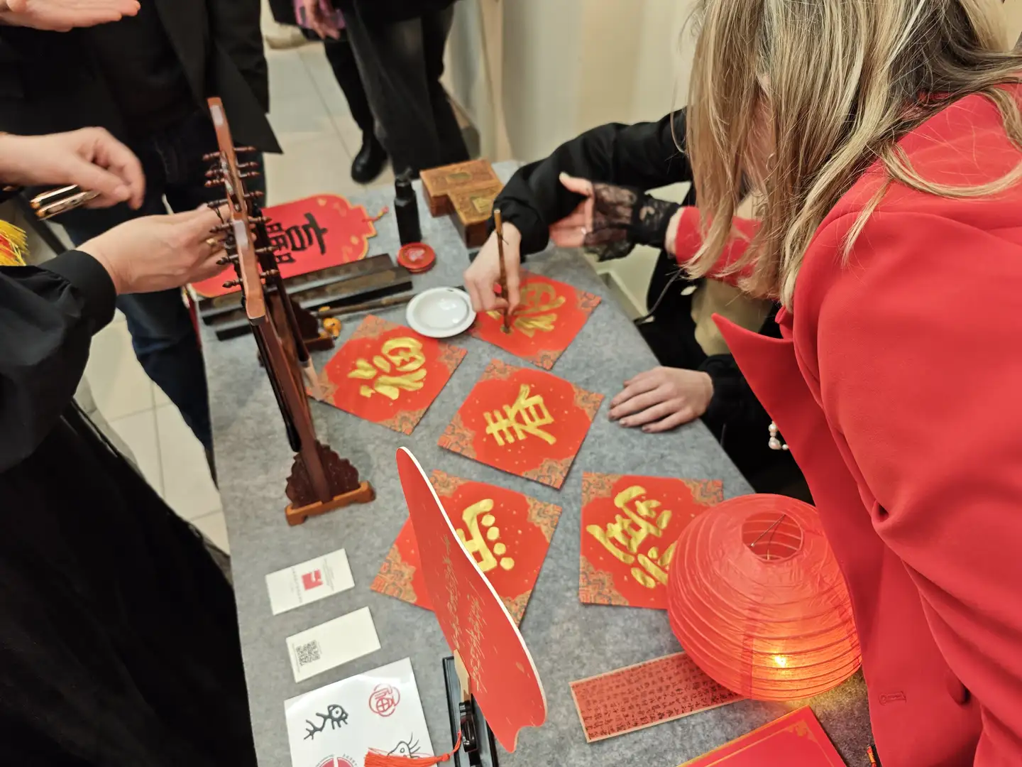 俄罗斯人写福字,圣彼得堡国立大学举办的春节庆祝活动 几个人正在一张灰色桌面上书写和展示红色的春联及菱形福字,有人正拿着毛笔在纸上写字。桌面上平铺着几张写有金色大字如“春”、“福”、“喜”的红色方块纸,旁边还放着一个亮着的红色圆形灯笼。左侧有一个木制的笔架和一些文房四宝用品,右下角有一个穿着红色外套的人背影。