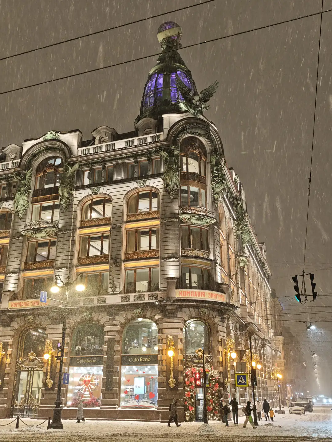 雪中的“书籍之家”,圣彼得堡 一座华丽的新艺术风格建筑矗立在雪夜的街角,外墙装饰着复杂的雕塑和浮雕,顶部有一个发光的紫色圆顶和一只展翅的鹰雕塑。建筑底层的橱窗明亮,招牌上写着“Cafe René”和俄文“КОМПАНИЯ ЭНГЕРЪ”,门口装饰着圣诞花环。天空中飘着大雪,街道和人行道上覆盖着积雪,行人穿着冬装在路灯下行走,远处有车辆行驶。