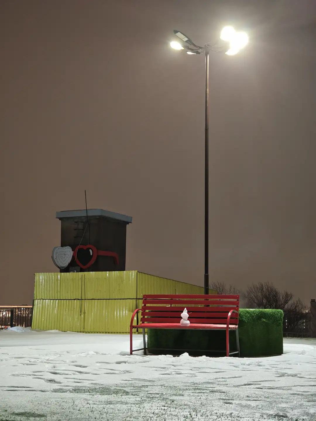 冬寂,观景阁楼,圣彼得堡 夜晚的雪地场景中,一盏高大的路灯发出明亮的白光,照亮了周围的区域。路灯下方有一张红色的长椅,长椅中间坐着一个微小的白色雪人,长椅背后是绿色的草皮状结构。左侧有一个黄色的波纹板围挡,上面立着一个深色的小房子结构,装饰着心形图案。