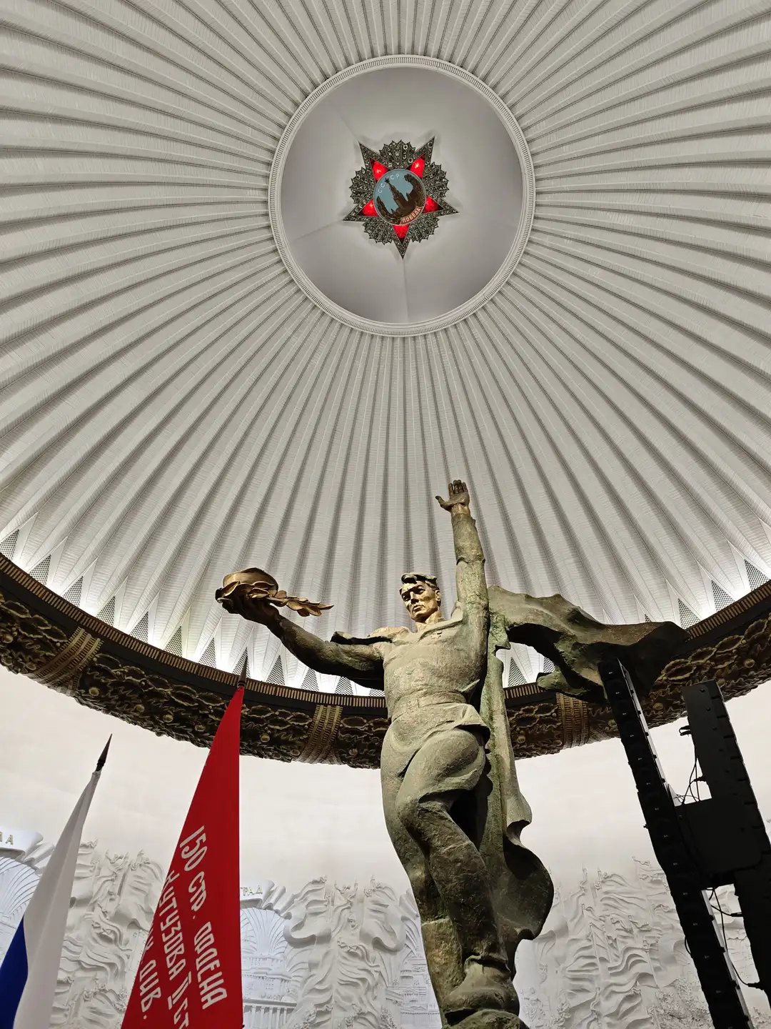 《胜利的战士》(«Солдат-победитель»),胜利博物馆,莫斯科 仰视视角下,一个巨大的白色放射状穹顶覆盖着空间,中心悬挂着一枚带有红星和地球图案的徽章。穹顶下方矗立着一座青铜色士兵雕像,他左手高举,右手托举着月桂枝花环,身披飘扬的披风。雕像后方是白色的浮雕墙壁,前景左侧有一面红色的旗帜,上面印有白色的西里尔字母文本。