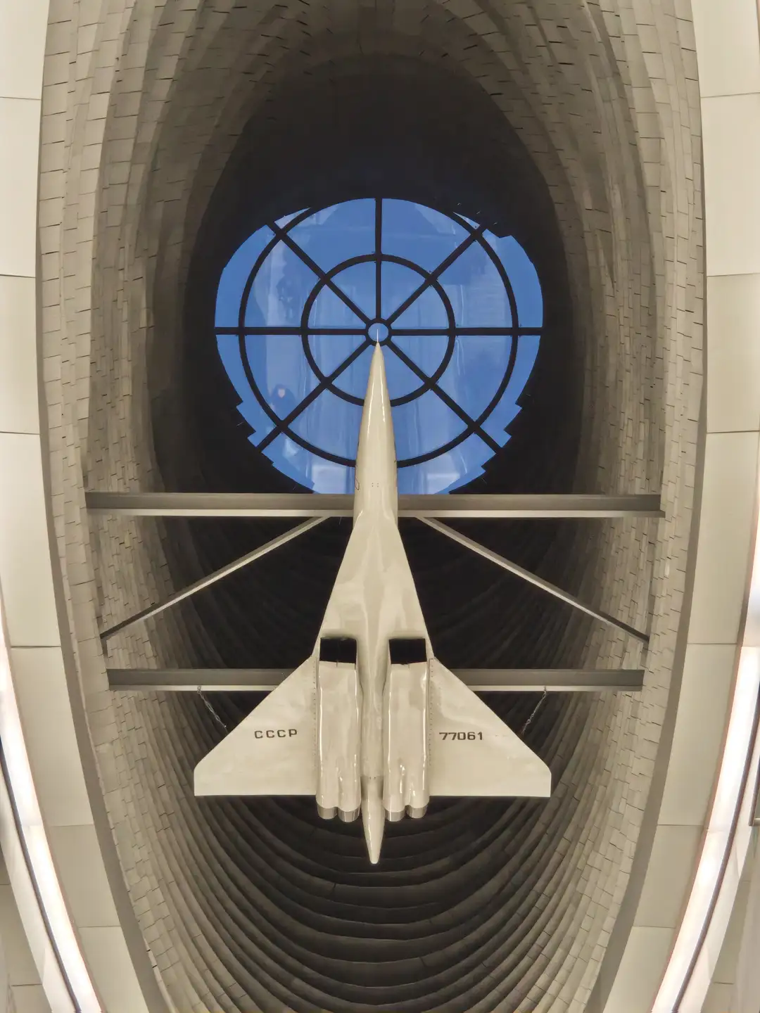 Tu-144D 飞机模型(虚构编号 CCCP-77061),地铁站“皮赫蒂诺”(«Пыхтино»),莫斯科 一架白色的超音速客机模型悬挂在巨大的拱形隧道结构中央,机身上印有“CCCP”和“77061”字样。飞机模型通过金属支架固定,正对着上方一个巨大的圆形蓝色窗户,窗户上有黑色的辐射状线条。周围的墙壁由层层叠叠的灰色砖块或板材构成,形成深邃的纵深感。