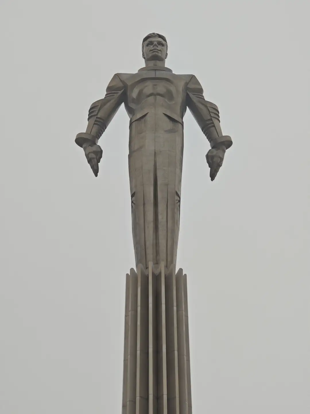 加加林纪念像,坐标 55.708311, 37.582539,莫斯科 一座高耸的灰色金属雕像展示了一位身穿宇航服的男性,他双臂自然下垂,目光凝视前方。雕像下半身逐渐演变为火箭喷射出的尾焰形状,由多根垂直的金属柱支撑。背景是灰白色的天空,雕像整体呈现出一种向上飞升的几何形态。