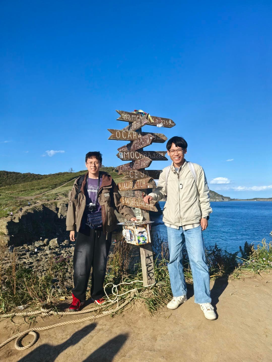 两个人站在海边,背景是蓝色的天空和海洋。他们身后有一个指示牌,上面写着不同城市的距离。左边的人穿着棕色外套和黑色裤子,右边的人穿着浅色夹克和蓝色牛仔裤。