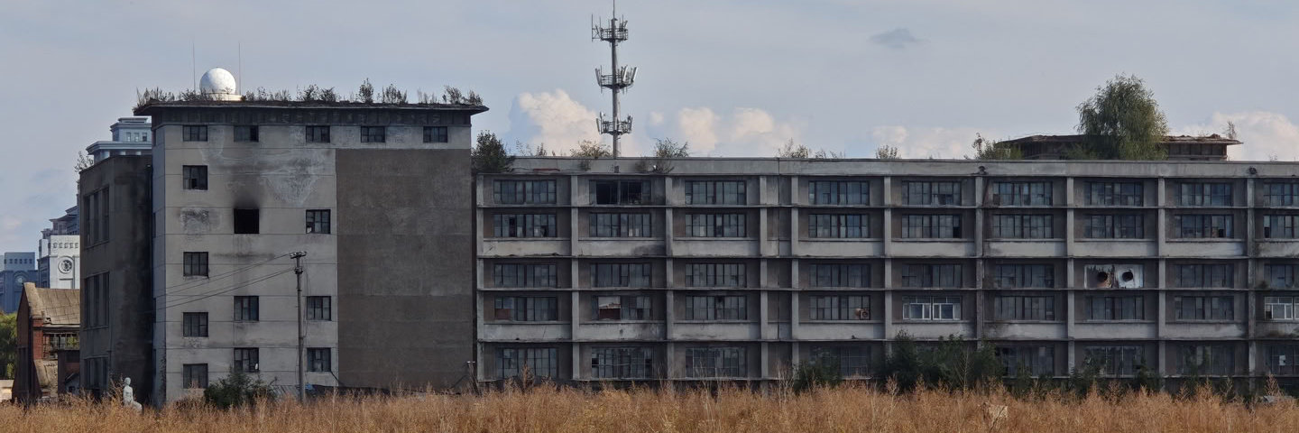 破败的建筑上杂草丛生,勾勒出秋日萧条的景象 一座长条形的灰色多层建筑横跨画面,窗户排列整齐但部分破损。建筑左侧连接着一栋较高的楼房,屋顶设有白色球状物体。建筑后方矗立着一座通信铁塔,前景是大片枯黄的杂草。