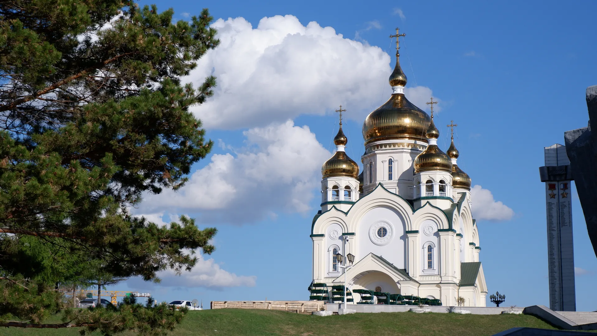 A white church topped with five golden domes and crosses. Lush pine trees stand to the left of the church against a background of blue sky and white clouds, with fences and vehicles in front.
