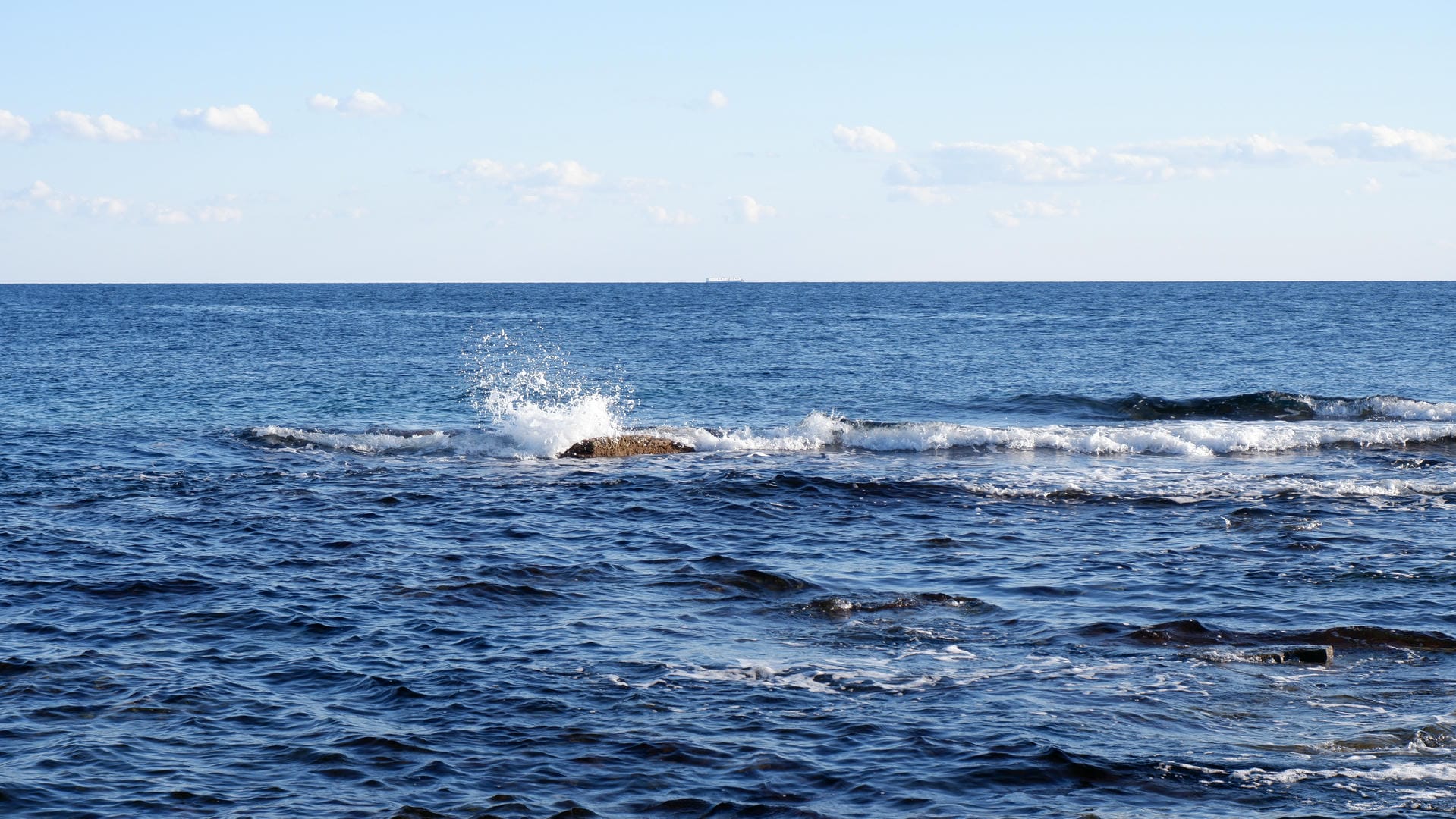Waves crashing against rocks and splashing white foam, with a few white clouds on the distant horizon.